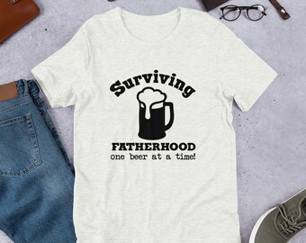 May include: A white t-shirt with a black graphic that says "Surviving Fatherhood one beer at a time!" and a beer mug with foam on top.