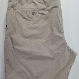 May include: Stone-coloured shorts with a buttoned back pocket and a button closure at the waist. The shorts are made of a lightweight fabric and appear to be unworn. The shorts are folded and laid flat on a white surface.