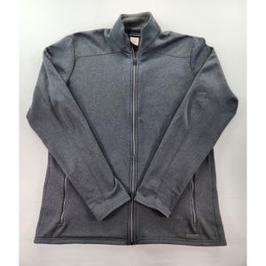 Puede incluir: Chaqueta gris con cremallera completa y cuello alzado. La chaqueta tiene dos bolsillos con cremallera y un pequeño logotipo en la parte inferior derecha. La chaqueta está hecha de un material suave y flexible.