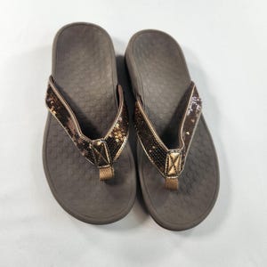 May include: A pair of brown flip-flops with sequined straps. The footbeds are a darker brown with a textured surface. The straps are embellished with bronze-colored sequins and a matching toe post.
