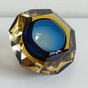 Könnte beinhalten: Ein facettierter Glasbriefbeschwerer mit einer tief bernsteinfarbenen Außenschicht mit geometrischen Schliffen und einer glatten, kreisförmigen, leuchtend blauen Mitte. Das Stück enthält ein kleines schwarzes Etikett mit dem weißen Text GNIN & BO.
