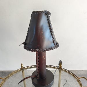 Può includere: Una lampada da tavolo artigianale con paralume e base in pelle marrone scuro. Il paralume è cucito lungo i bordi e la lampada poggia su una base rotonda. La lampada è su un tavolo con piano in vetro e cornice in ottone.