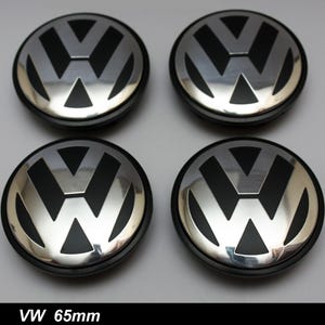 Könnte beinhalten: Vier silberne und schwarze VW-Radnabenabdeckungen. Jede Kappe zeigt das VW-Logo in Silber auf schwarzem Hintergrund. Die Kappen sind rund mit einem schwarzen Außenring. Der Text "VW 65mm" steht unten.