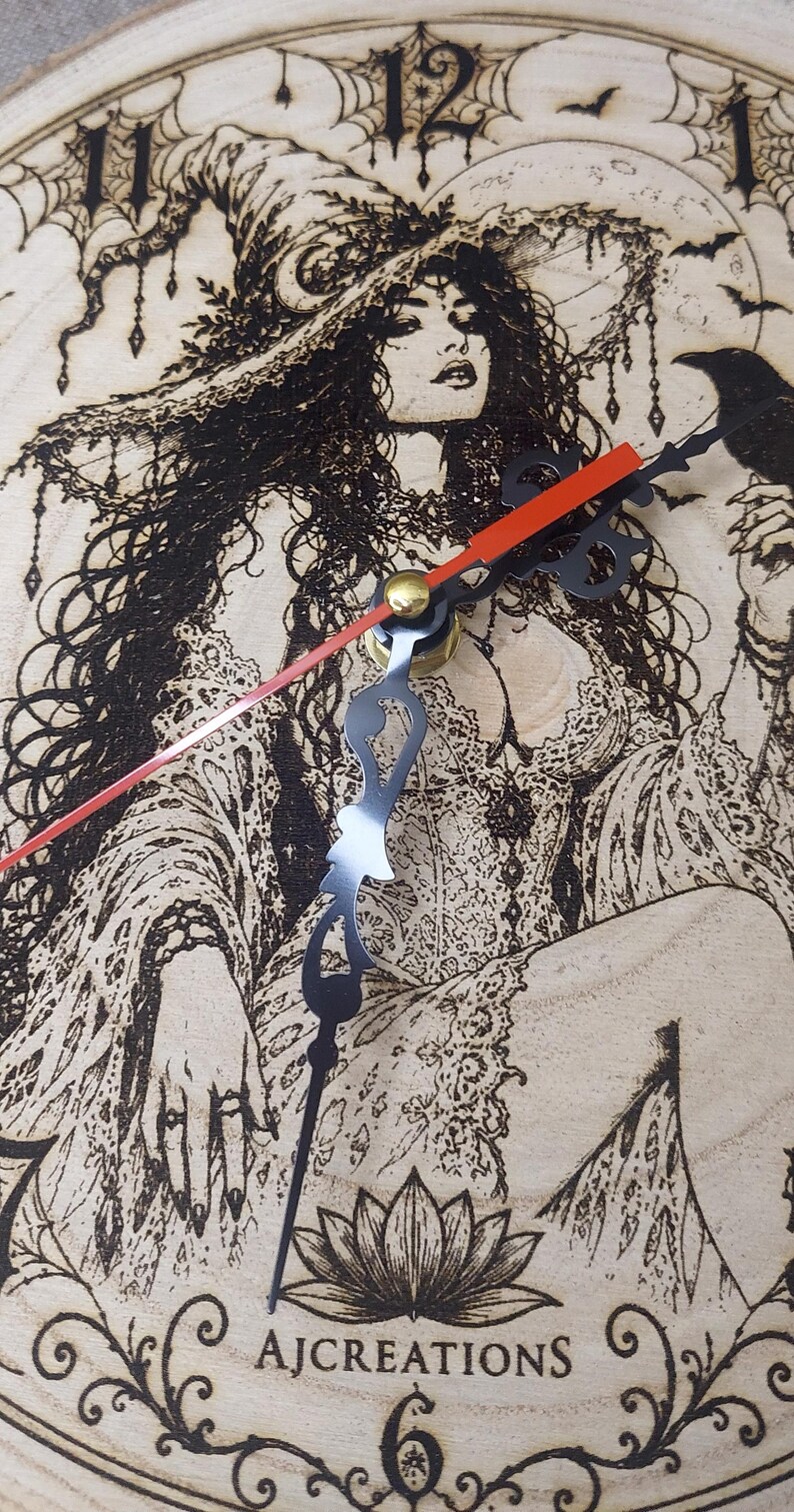 Pu&ograve; includere: Un orologio da parete rotondo in legno con un'illustrazione pirografata di una figura con un cappello a tesa larga, un corvo, ragnatele e un fiore di loto. Il quadrante presenta numeri neri, lancette nere decorate e una sottile lancetta dei secondi rossa. Il testo AJCREATIONS &egrave; stampato in basso.