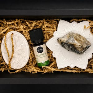 Op de afbeelding: Een zwarte geschenkdoos met een wit ovaal keramisch ornament met een gouden koord, een klein glazen flesje Citronnelle etherische olie met een zwarte dop, en een wit stervormig keramisch schaaltje met een ruwe, grijs-witte minerale steen.
