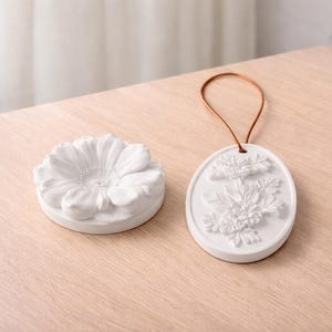 May include: Two white ceramic decorative items on a light wood surface. One is a flower-shaped dish, the other is an oval ornament with a floral design and a brown leather loop. Suitable for home decor.