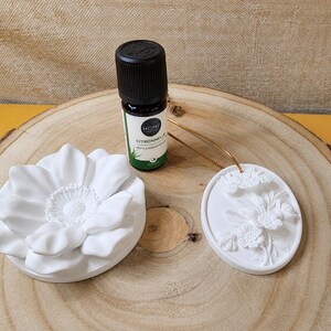 Peut inclure: Un petit flacon marron foncé d'huile essentielle Homi Citronnelle à côté d'un diffuseur en céramique blanche en forme de fleur et d'une décoration suspendue ovale en céramique blanche avec un relief floral et un cordon doré.