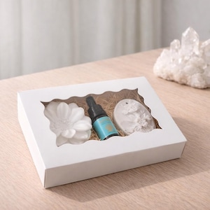 Può includere: Una confezione regalo bianca con una finestra trasparente, contenente un oggetto bianco a forma di fiore, una piccola bottiglia blu e un oggetto bianco ovale. La scatola è su una superficie di legno marrone chiaro, con un gruppo di cristalli bianchi sullo sfondo.