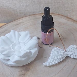 May include: A white sculpted flower diffuser, a small black dropper bottle with a pink label, and a white pair of angel wings ornament with a thin gold hanging cord. The items are arranged together on a circular light-colored wooden base.