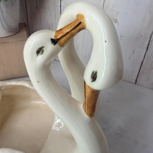 May include: A cream-colored ceramic swan planter featuring two swan necks that curve toward each other to form a heart shape. The swans have hand-painted orange beaks with black details and small green eyes, finished with a glossy glaze.