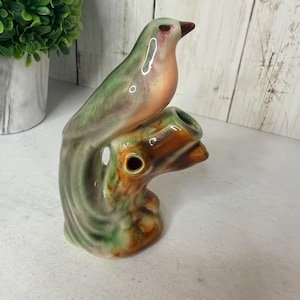 Pode incluir: Uma estatueta de cerâmica de um pássaro pousado num ramo de árvore, com um esmalte brilhante em tons misturados de verde sálvia, laranja suave e castanho. O pássaro tem um bico castanho escuro e o ramo inclui uma abertura oca na lateral.