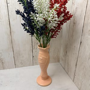 May include: A peach-colored ceramic vase with a flared rim and textured leaf-like pattern holds a bouquet of artificial flowers in deep blue, white, and bright red, arranged to mimic the vertical stripes of a national flag.