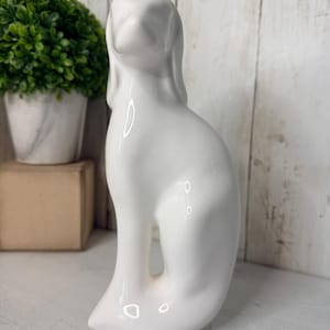 May include: A smooth, white ceramic figurine of a dog in a seated position. The sculpture features a minimalist, stylized form with long, drooping ears and a sleek, glossy finish that reflects light, capturing the canine's upright posture.