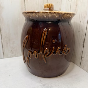 May include: A dark brown ceramic cookie jar with a glossy finish and a matching lid. The word Cookies is embossed in a cursive script on the front of the jar. The rim and the lid feature a lighter brown, drip-style glaze pattern.
