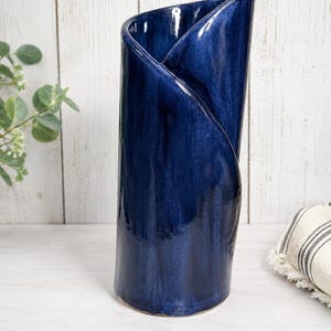 May include: A tall, cylindrical ceramic vase with a deep navy blue glossy glaze. The vase features a unique overlapping, folded design at the top rim, creating a diagonal opening. The surface has subtle variations in color depth and texture.