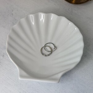 May include: A white ceramic trinket dish shaped like a scallop shell with a ribbed surface. Two silver-toned rings are resting in the center of the dish. One ring features a thin band with small embedded stones, and the other has a single stone setting.