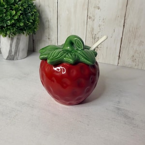 May include: A red ceramic strawberry-shaped jar with a green leaf lid and a small white spoon. The jar features a textured surface with dimples mimicking strawberry seeds. The lid has a sculpted stem handle and detailed leaf patterns.