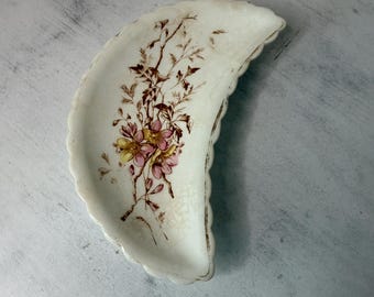 May include: A crescent-shaped ceramic dish with a scalloped edge, featuring a cream-colored base and a detailed botanical illustration of pink and yellow flowers with brown stems and leaves centered on the interior surface of the curved plate.
