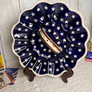 May include: A round ceramic egg plate with a scalloped edge, featuring a deep blue glaze decorated with numerous small white stars. The plate includes a central handle with brown and white stripes and multiple circular indentations for eggs.