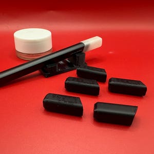 May include: A selection of black 3D-printed accessories, including small, rectangular pieces and a long, cylindrical object with a white tip. A small, white-lidded jar is also present. The items are displayed on a solid red surface.