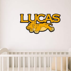 May include: A wall-mounted sign featuring the name LUCAS in bold yellow capital letters with a black outline, positioned above a graphic of a sleeping yellow lion cub with black outlines, displayed on a plain white wall above a white crib.