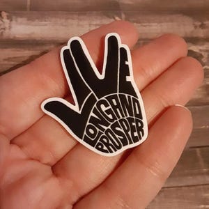 May include: A black and white sticker in the shape of the Vulcan salute hand gesture. The words "Live Long and Prosper" are printed in white within the hand shape. The sticker is held in a hand.