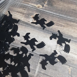 May include: Black witch-shaped confetti scattered on a wooden surface. The confetti pieces are cut-out silhouettes of witches riding brooms, ideal for Halloween decorations. The dark color of the confetti contrasts with the light wood grain.