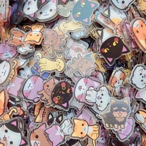 May include: A large collection of small, clear acrylic charms featuring colorful, cartoon-style illustrations of various cats and dogs with expressive faces, layered together in a dense, overlapping pile.