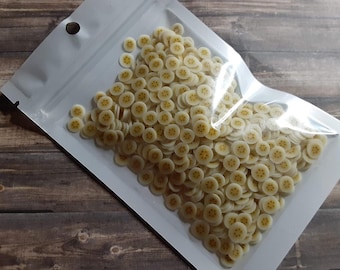 May include: A clear plastic bag filled with numerous small, round, banana-shaped craft embellishments. Each piece is off-white with a yellow center, resembling a sliced banana. The bag is on a dark brown wooden surface.
