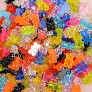 Puede incluir: Una colección de coloridos abalorios de ositos de goma en varios colores, incluyendo naranja, amarillo, rosa, azul y negro. Cada abalorio tiene un pequeño lazo plateado para sujetarlo a joyas u otras manualidades. Los abalorios están hechos de un material translúcido.
