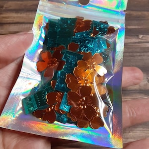 May include: A small, clear, iridescent resealable plastic pouch containing a collection of loose, metallic-finish craft embellishments. The contents consist of shiny copper-colored flower-shaped sequins and teal-colored rectangular pieces with embossed patterns.