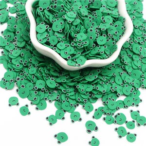 May include: A white bowl overflowing with green frog-shaped polymer clay sprinkles. The sprinkles have black eyes and a smiling mouth. The frog sprinkles are scattered around the bowl on a white surface.