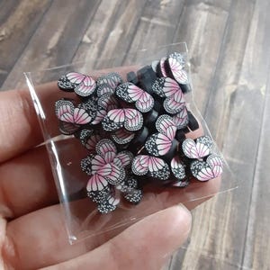 May include: A clear plastic bag filled with small, butterfly-shaped polymer clay pieces. The butterflies have pink and white wings with black detailing. The clay pieces are stacked together, creating a colorful and detailed pattern.