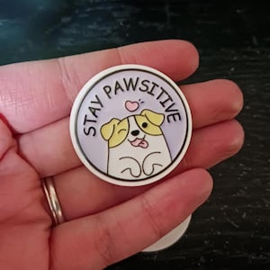 May include: A round, white and lavender pin with the words "STAY PAWSITIVE" encircling a cartoon dog illustration. The dog has yellow ears, a pink tongue, and a small pink heart above its head. The pin is held in a hand.