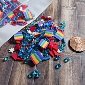 May include: A collection of colorful polymer clay sprinkles and blue iridescent beads are scattered on a wooden surface. The sprinkles include rainbow-colored squares, red triangles, and white snowflake shapes. A penny is included for size reference.