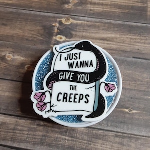 May include: A round, white pin with a blue glitter background. The pin features a black snake wrapped around a sign that reads "I JUST WANNA GIVE YOU THE CREEPS." Pink flowers are on the left side of the sign.