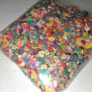 May include: A clear plastic bag filled with a variety of small, colorful polymer clay slices. The assortment includes shapes of brown bears, yellow crescent moons, rainbows, green frogs, and red lips in a dense, multi-colored pile.