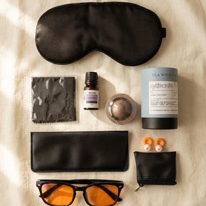 May include: A collection of sleep essentials, including a black eye mask, glasses with orange lenses, earplugs, a small bottle of lavender essential oil, and a tea canister labeled "Nighttime Soother". The items are arranged on a neutral-toned surface.