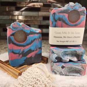 May include: Artisan soap bars with a swirl of pink, blue, and black. One bar is on a wooden soap dish, and another is stacked with two more. The soap has a circular cutout. The label reads "Goats Milk & Silk Soap, Houston, We Have a Bubble."