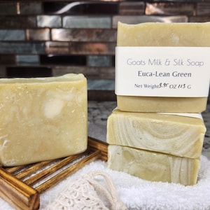 May include: Several bars of Euca-Lean Green goat milk and silk soap. The soap is a light green color with a marbled design. One bar rests on a wooden soap dish, while others are stacked on a white towel. The label reads "Goats Milk & Silk Soap".
