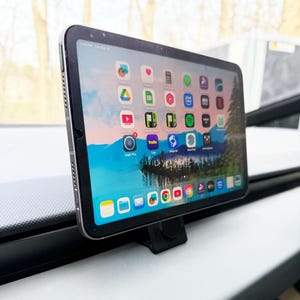 Peut inclure: Une tablette noire affichant diverses icônes d'applications, montée sur un support noir. La tablette est posée sur une surface blanche. L'arrière-plan est flou.