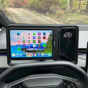 May include: Dashboard mount holding a tablet and a smartphone. The tablet displays app icons on a blue and green background. The smartphone shows the time as 9:57. The steering wheel is in the foreground.