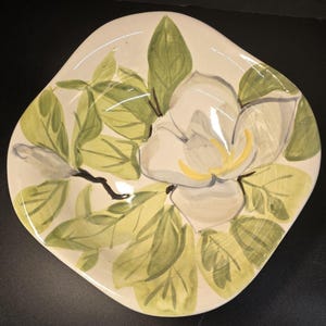 Puede incluir: Un plato de cerámica de color crema, de forma cuadrada, con un diseño de flor de magnolia pintado a mano. La flor es blanca con un centro amarillo, rodeada de hojas verdes y una rama marrón oscuro. El plato tiene un borde ligeramente irregular.