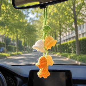May include: A handmade crochet car charm featuring a cluster of flower-shaped ornaments. The charm has a green stem and includes white, yellow, and orange flowers. The charm hangs from the rearview mirror, with a blurred street and trees in the background.
