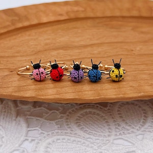 Könnte beinhalten: Fünf verstellbare Ringe aus goldfarbenem Metall, die jeweils mit einem kleinen, gehäkelten Marienkäfer verziert sind. Die Marienkäfer sind rosa, rot, lila, blau und gelb und haben schwarze Faden-Details für Kopf, Beine und Punkte.