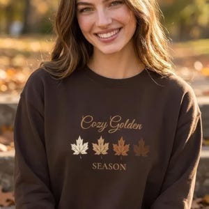 May include: A dark brown long-sleeve sweatshirt featuring the text Cozy Golden SEASON in a light brown font. Below the text Cozy Golden, there is a horizontal row of four maple leaf illustrations in varying shades of cream, tan, brown, and dark brown.