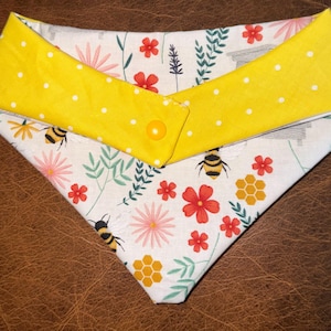 Puede incluir: Bandana para perros amarilla y blanca con un diseño de abejas y flores. La bandana tiene un cuello amarillo con lunares blancos y un cierre de botón amarillo. La tela principal presenta abejas, flores y diseños de panal.
