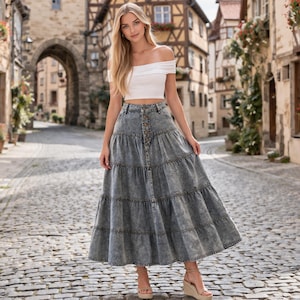 May include: A woman in a white crop top and a long, tiered denim skirt with a button-down front. The skirt has a faded wash. She wears wedge sandals and stands on a cobbled street, with a historic building backdrop.