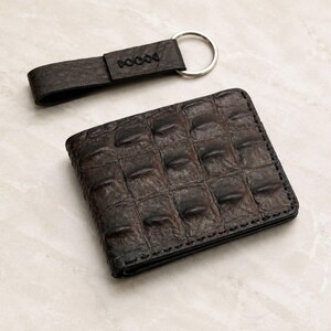 Peut inclure: Un ensemble portefeuille et porte-clés en cuir marron. Le portefeuille présente un motif gaufré texturé et des coutures noires. Le porte-clés a un anneau argenté et le mot "DOOOO" en relief sur la lanière en cuir. L'ensemble est présenté sur une surface claire.