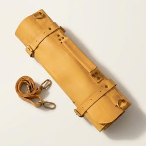 Könnte beinhalten: Eine aufgerollte, hellbraune Leder-Messertasche mit passendem Schulterriemen. Die Rolle wird mit mehreren Riemen und Metallbeschlägen gesichert. Der Riemen hat Metallverschlüsse. Auf weißem Hintergrund.
